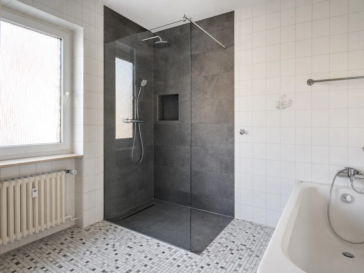 Teilsanierte Dusche mit dunklen Fliesen neben erhaltenen weißen Wandfliesen und Badewanne.